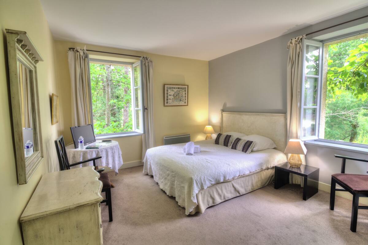 A bedroom with a bed, bedside tables, a desk, a chair, and large windows with curtains.