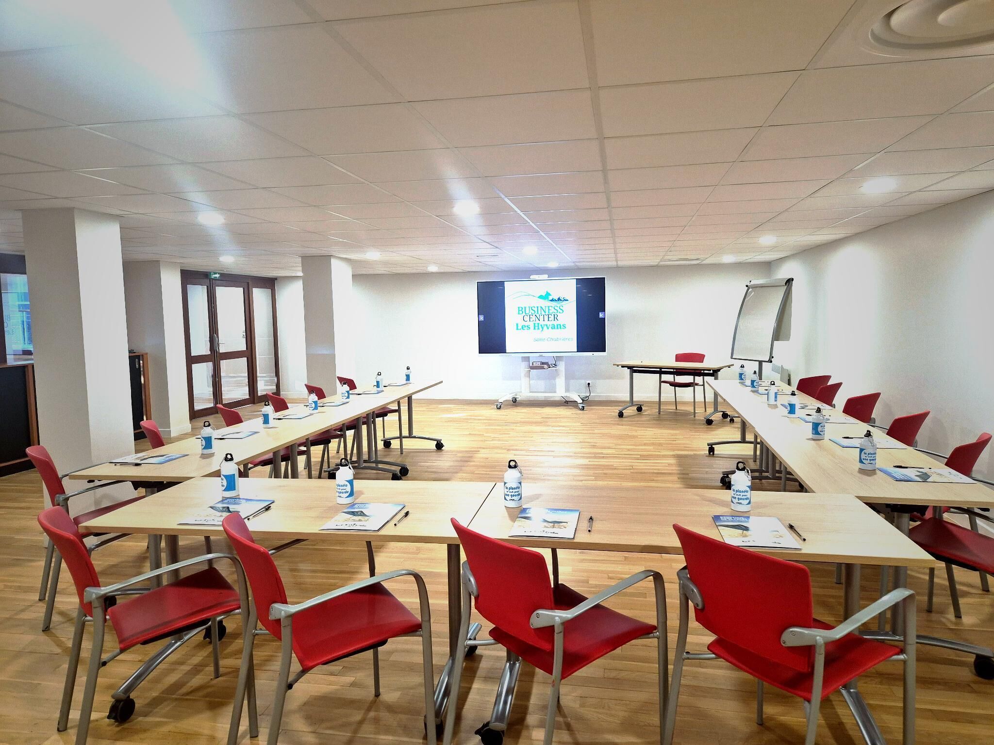 Een vergaderzaal met een lange tafel, rode stoelen, een projectiescherm en een presentatiescherm.