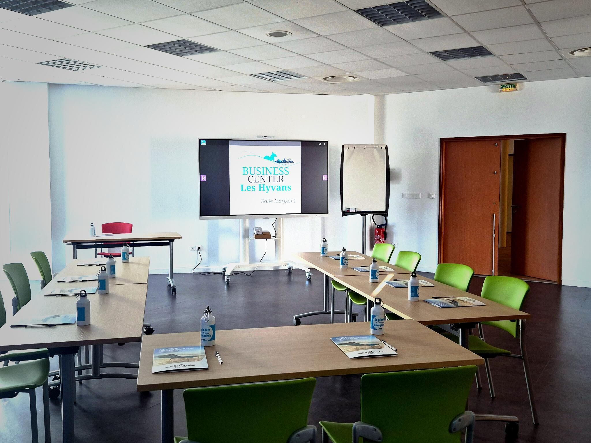 Una sala conferenze con un grande schermo che mostra 'BUSINESS CENTER Les Hyvans', una lavagna e sedie verdi disposte attorno a un lungo tavolo con bottiglie d'acqua e documenti.