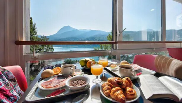 A breakfast table with food and drinks, a book, and a view of mountains through a window. - Village Club les Hyvans (Chorges)
