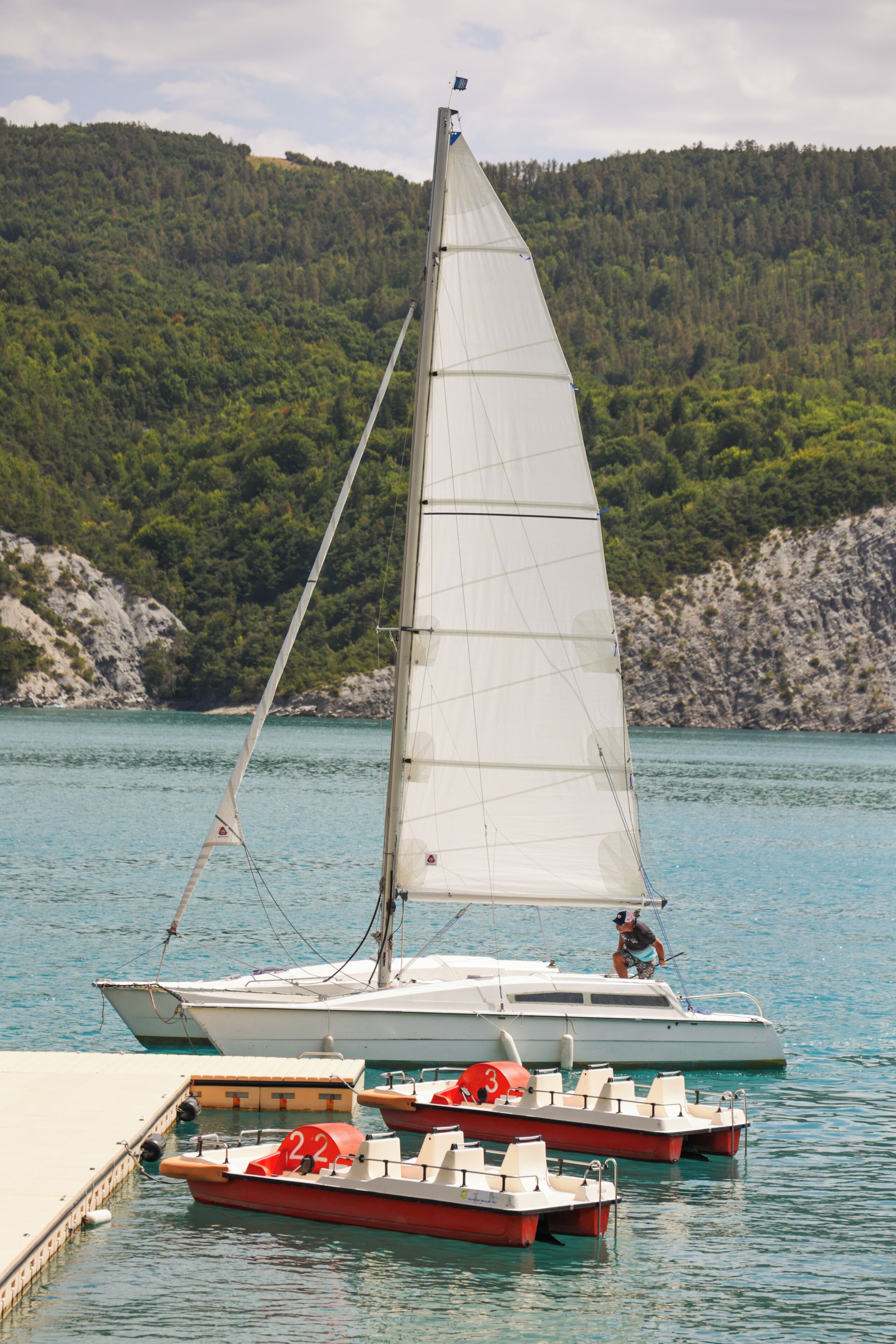 A sailboat docked at a pier with a forested hill in the background.