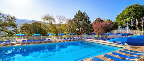 Una piscina con sillas reclinables azules y sombrillas, rodeada de árboles y montañas al fondo.