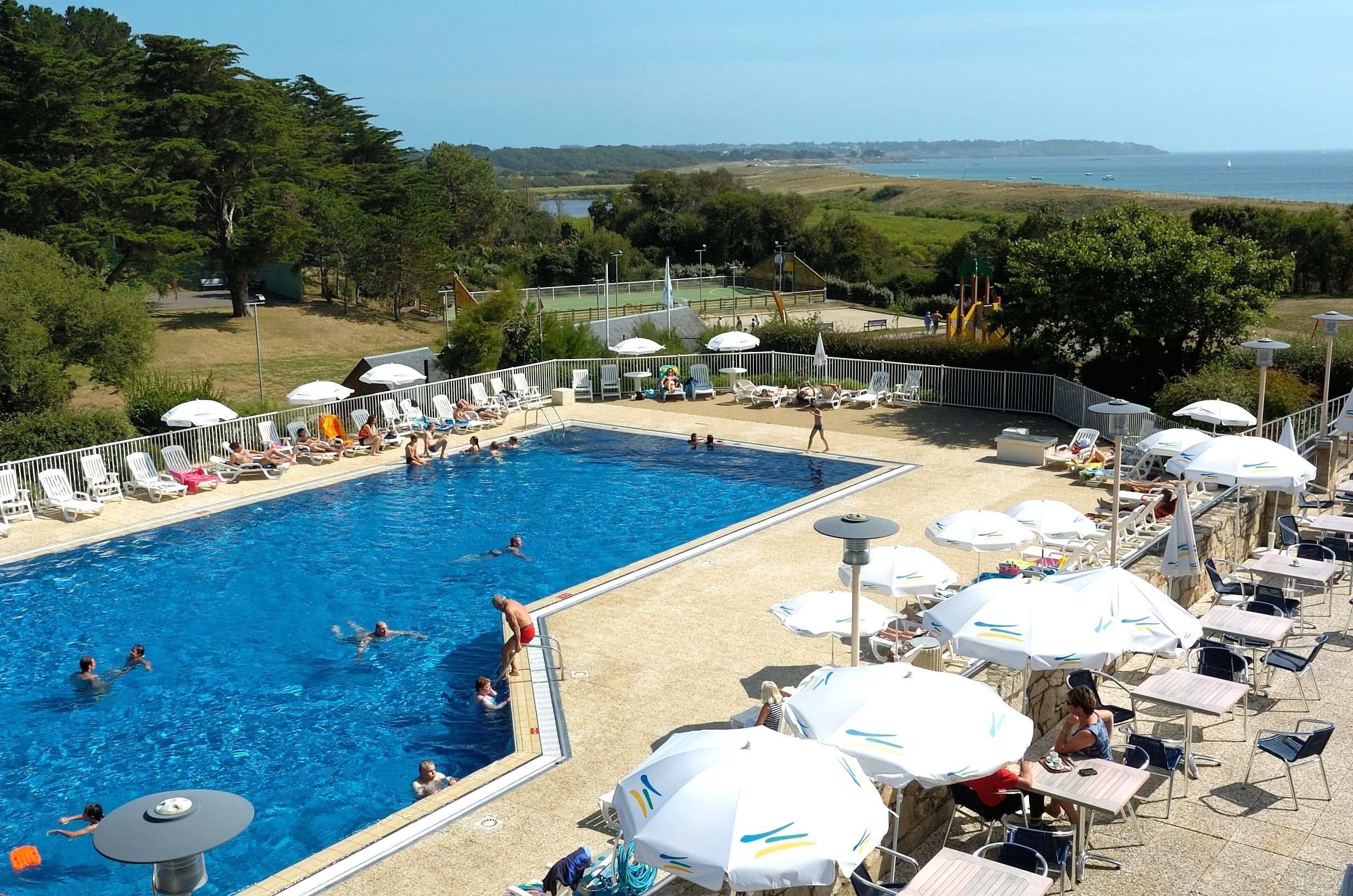 A swimming pool with people swimming and sunbathing, surrounded by umbrellas and lounge chairs.