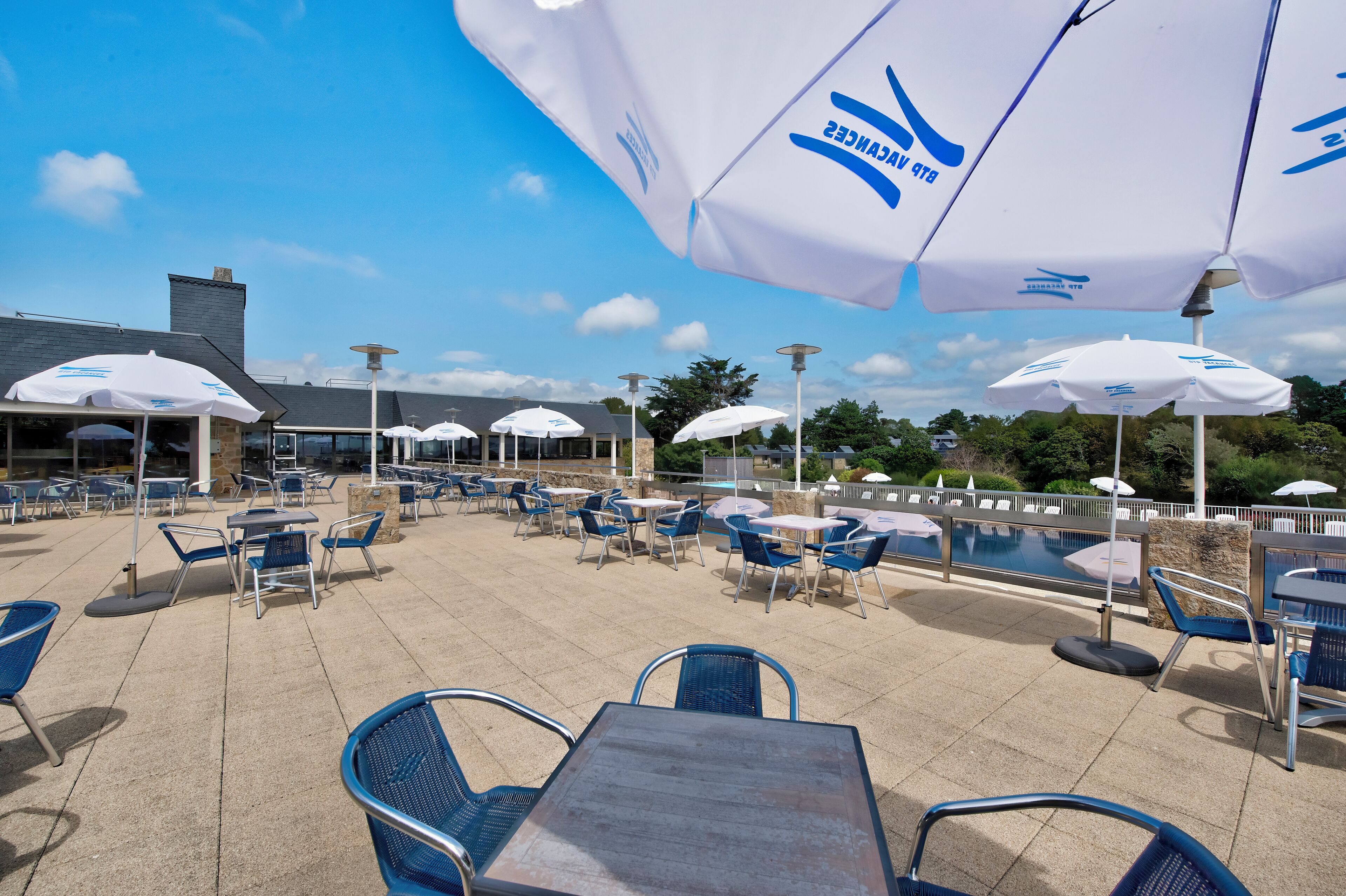 Een terras buiten met tafels, stoelen, parasols en een zwembad.