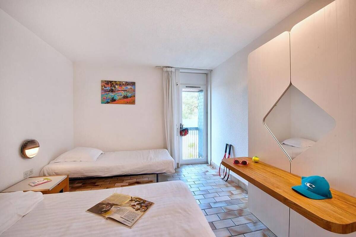 A compact hotel room with two single beds, a wooden side table with a cap, a small desk with a lamp, and a painting on the wall.