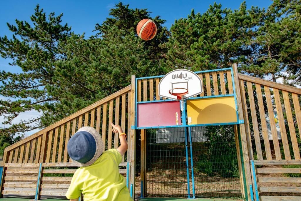 Ett barn som spelar basketboll på en utomhusplan med ett trästaket och träd i bakgrunden.