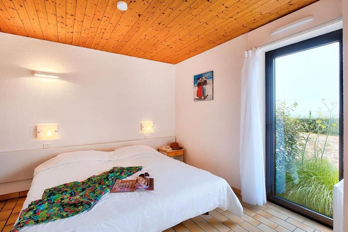 A bedroom with a wooden ceiling, a large bed with white bedding, two bedside lamps, a nightstand, a painting, and a view through a large window.