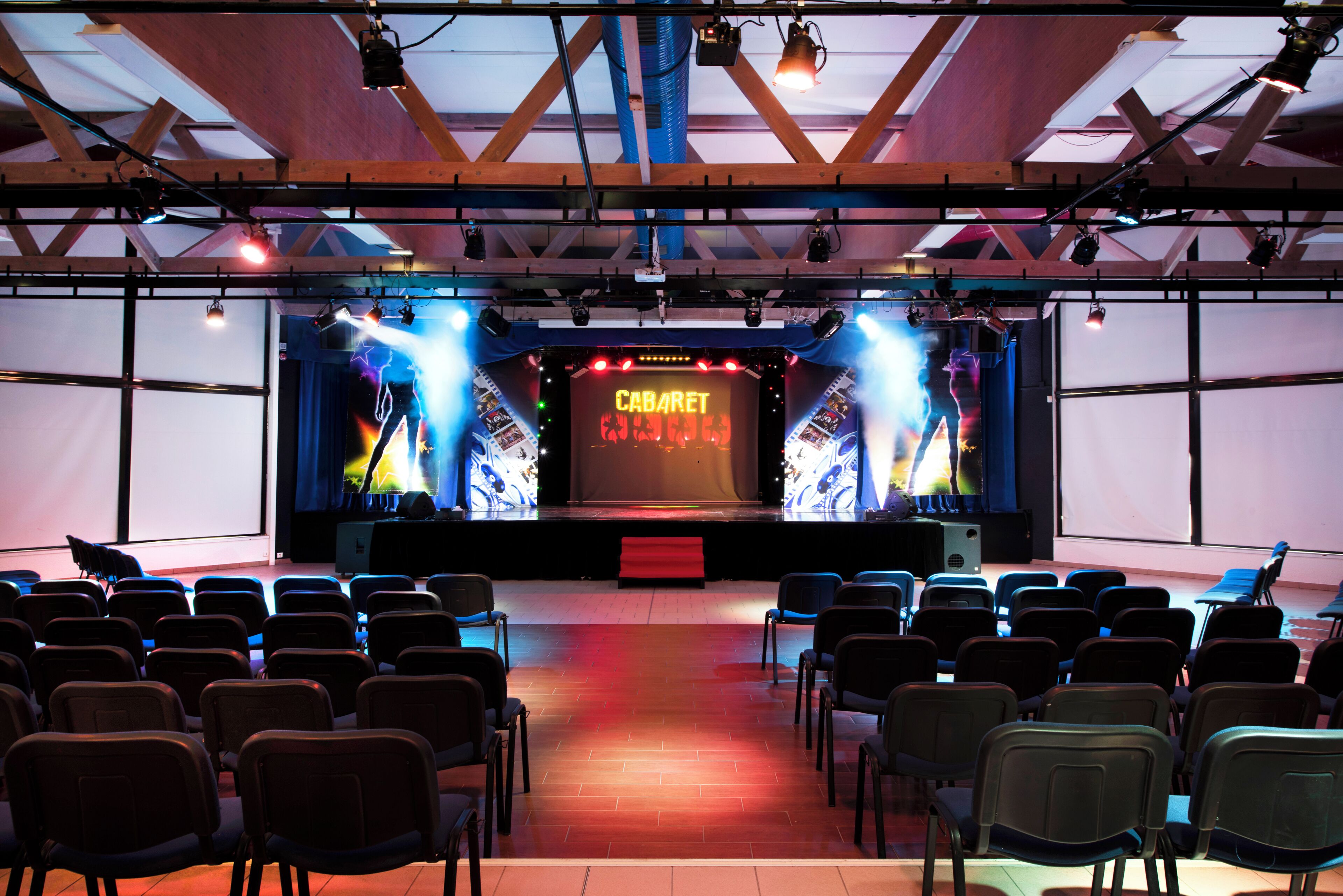 Ein Auditorioum mit Reihen von Stühlen, die auf eine Bühne zeigen, auf der ein großer Bildschirm 'CABARET' zeigt.
