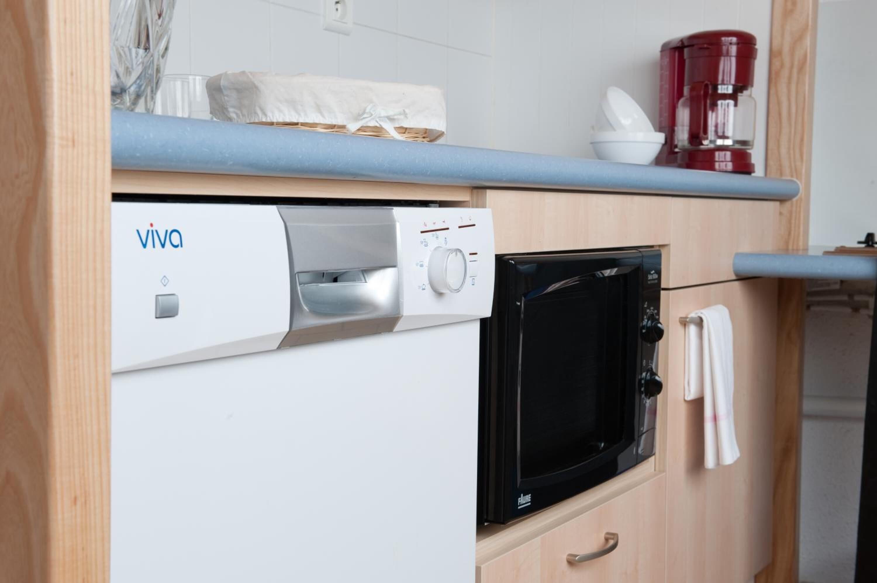 A compact kitchen with a white dishwasher, a black microwave, and a red coffee maker on the countertop.