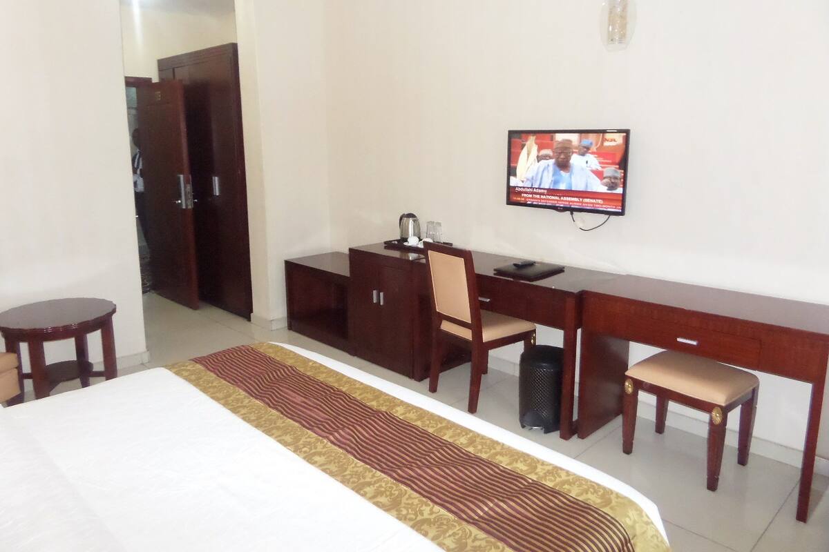 Quarto de hotel com cama, escrivaninha, cadeira, televisão e guarda-roupa.