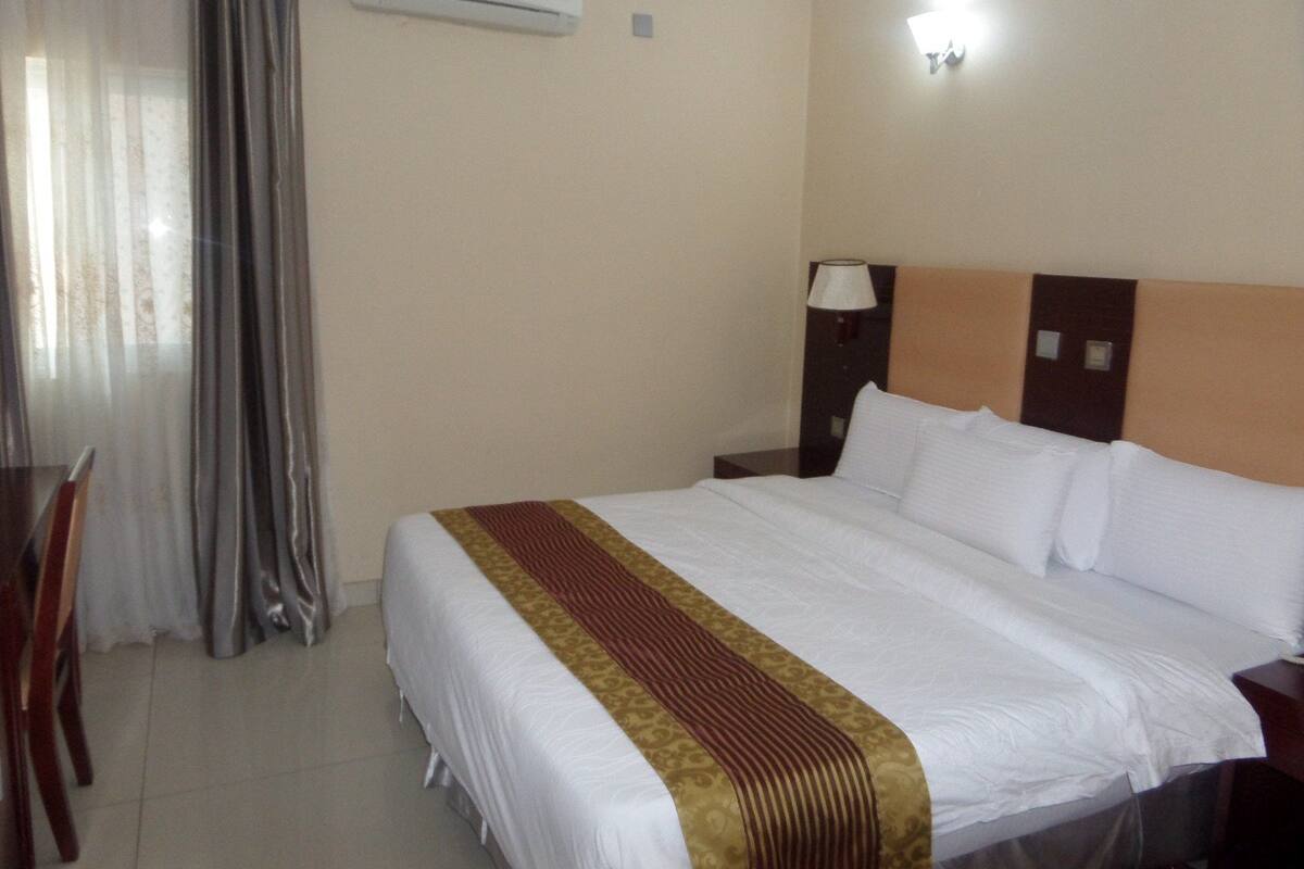 Quarto de hotel com cama, mesa de cabeceira, luminária, aparelho de ar condicionado e janela com cortinas.
