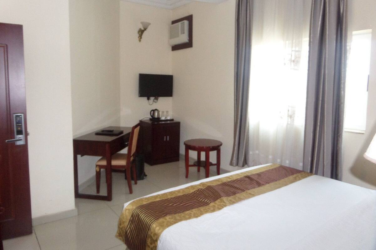 Quarto de hotel com cama, escrivaninha, cadeira, televisão e uma pequena mesa.