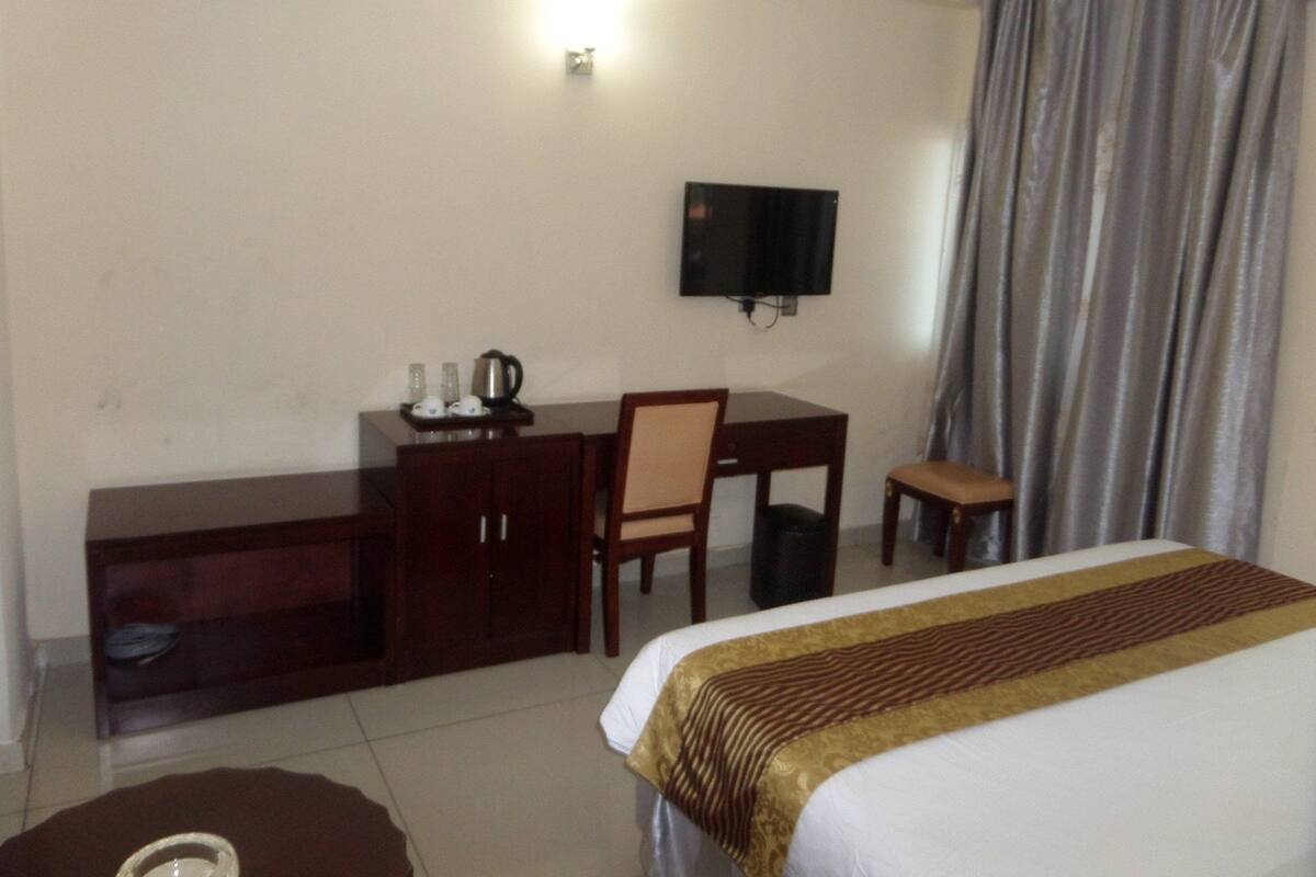 Quarto de hotel com cama, escrivaninha, cadeira, televisão e uma pequena mesa com chaleira e chávenas.