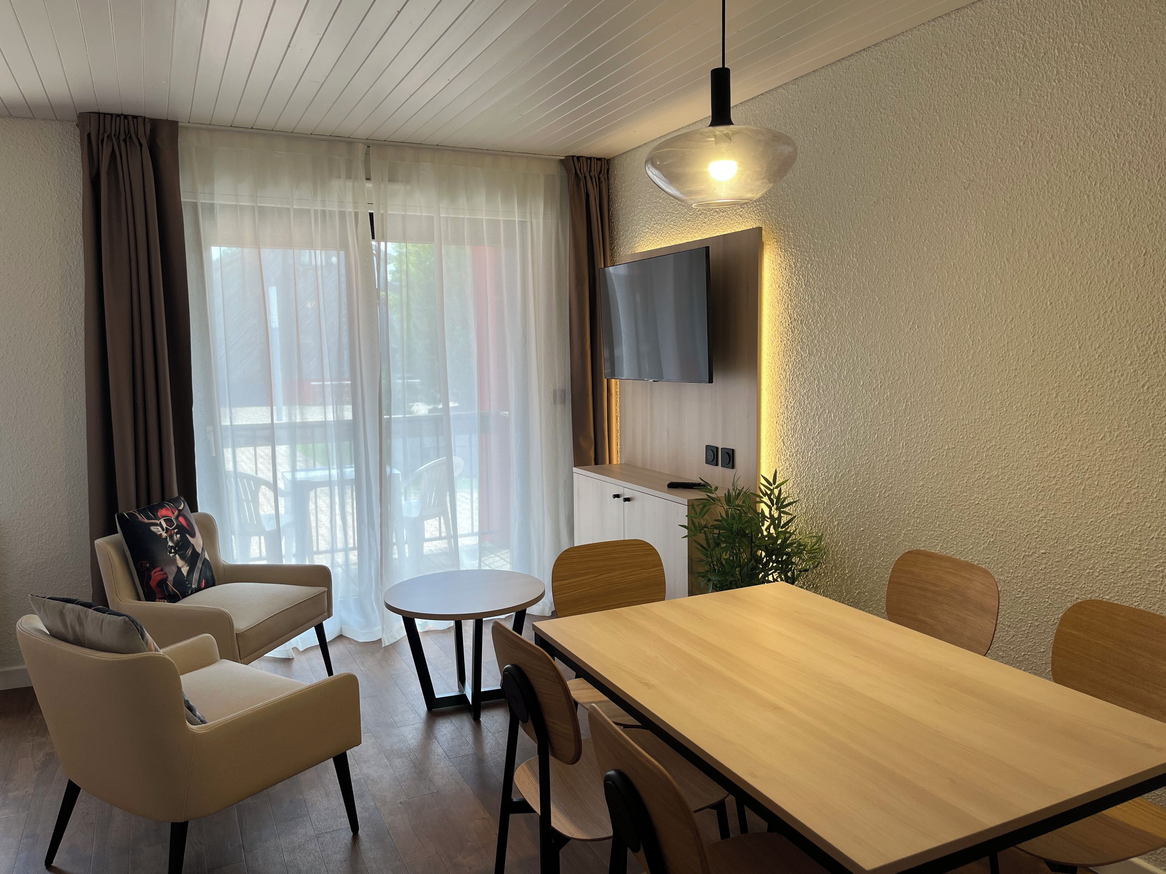 A modern living room with a wooden dining table, comfortable chairs, a flat-screen TV mounted on the wall, and a sliding glass door leading to an outdoor area.