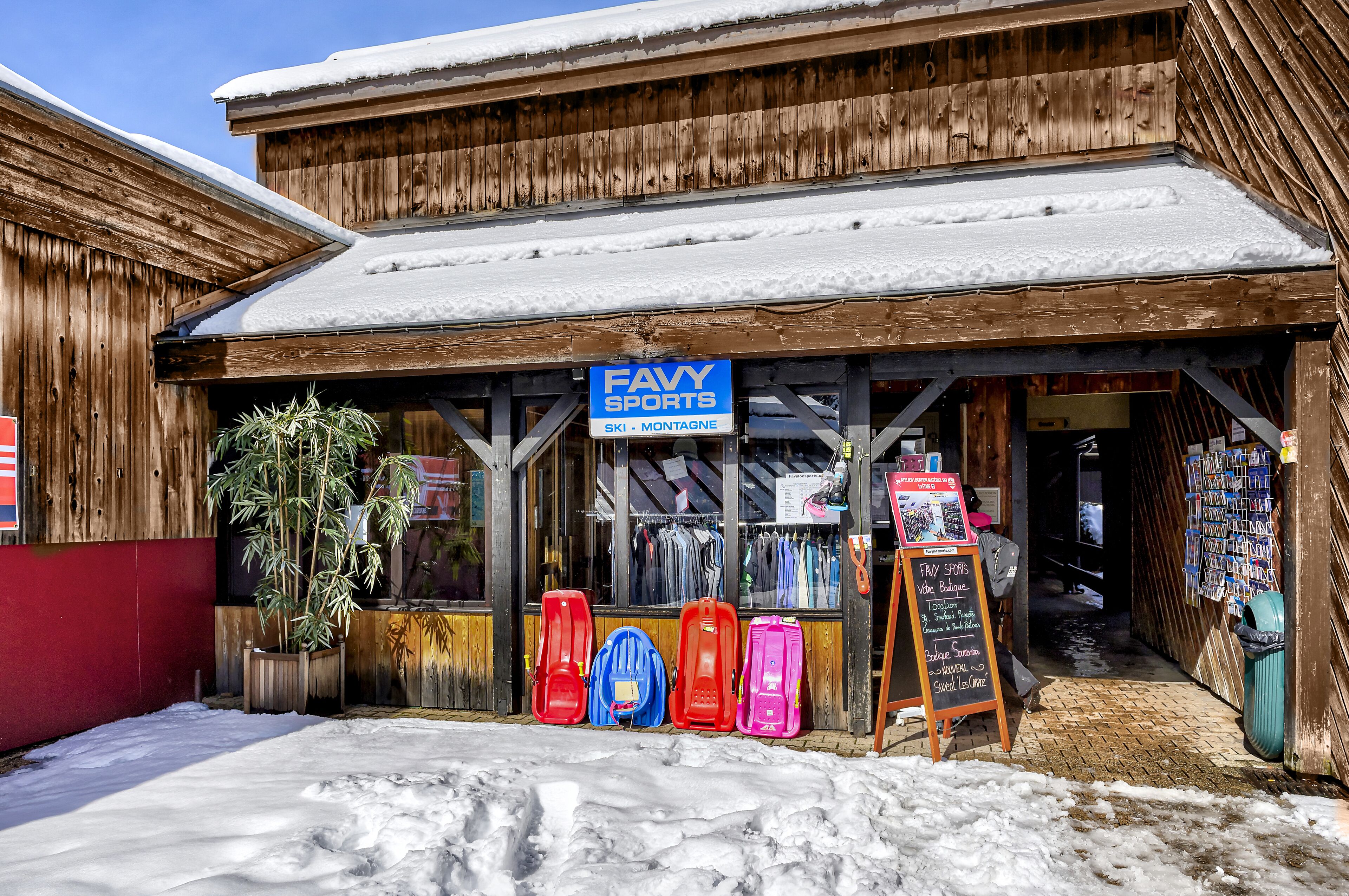 A wooden building with a sign for Favy Sports, ski and snowboard equipment, and a signboard listing prices.