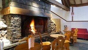 Una chimenea de piedra con fuego, rodeada de mesas y sillas de madera, en un ambiente interior acogedor.
