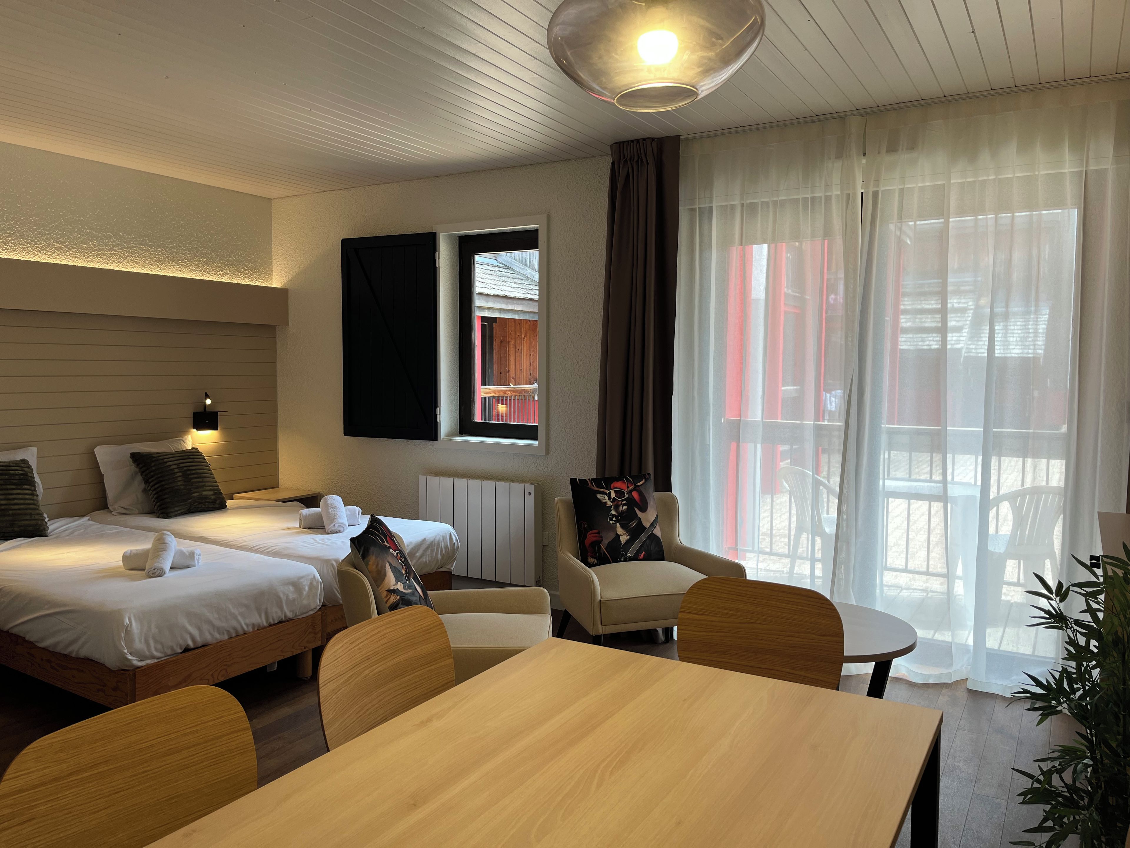 Ett hotellrum med två sängar, ett matbord, en stol, en soffa, ett stort fönster med gardiner och en taklampa.