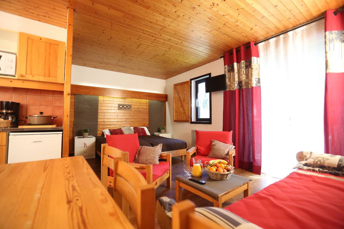 Een knus interieur met een houten plafond, een bed met rode beddengoed, een eettafel en een keukenhoek met een koffiezetapparaat.