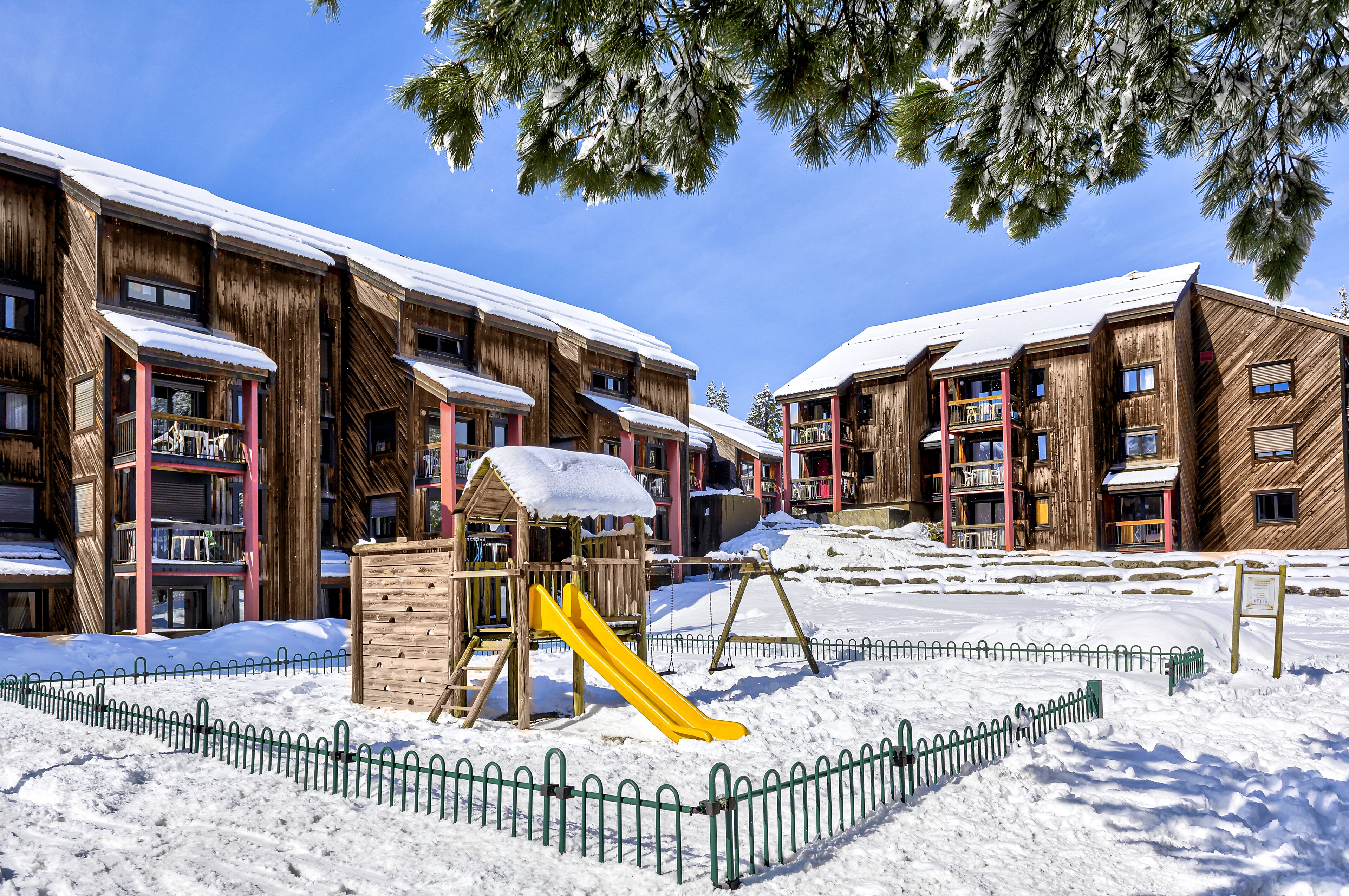 A snowy outdoor area with wooden buildings, a playground with a yellow slide, and a fenced play area.