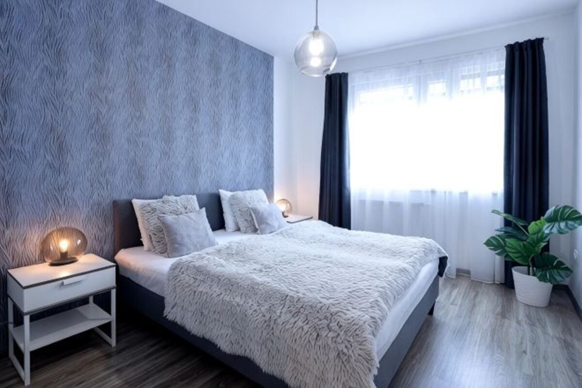 A modern bedroom with a bed, bedside tables, a hanging light fixture, a window with curtains, and a potted plant.