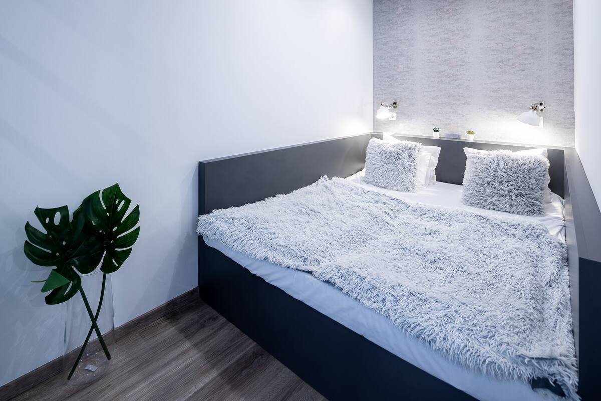 A modern bedroom with a dark headboard, white fluffy bedding, and a large green plant.