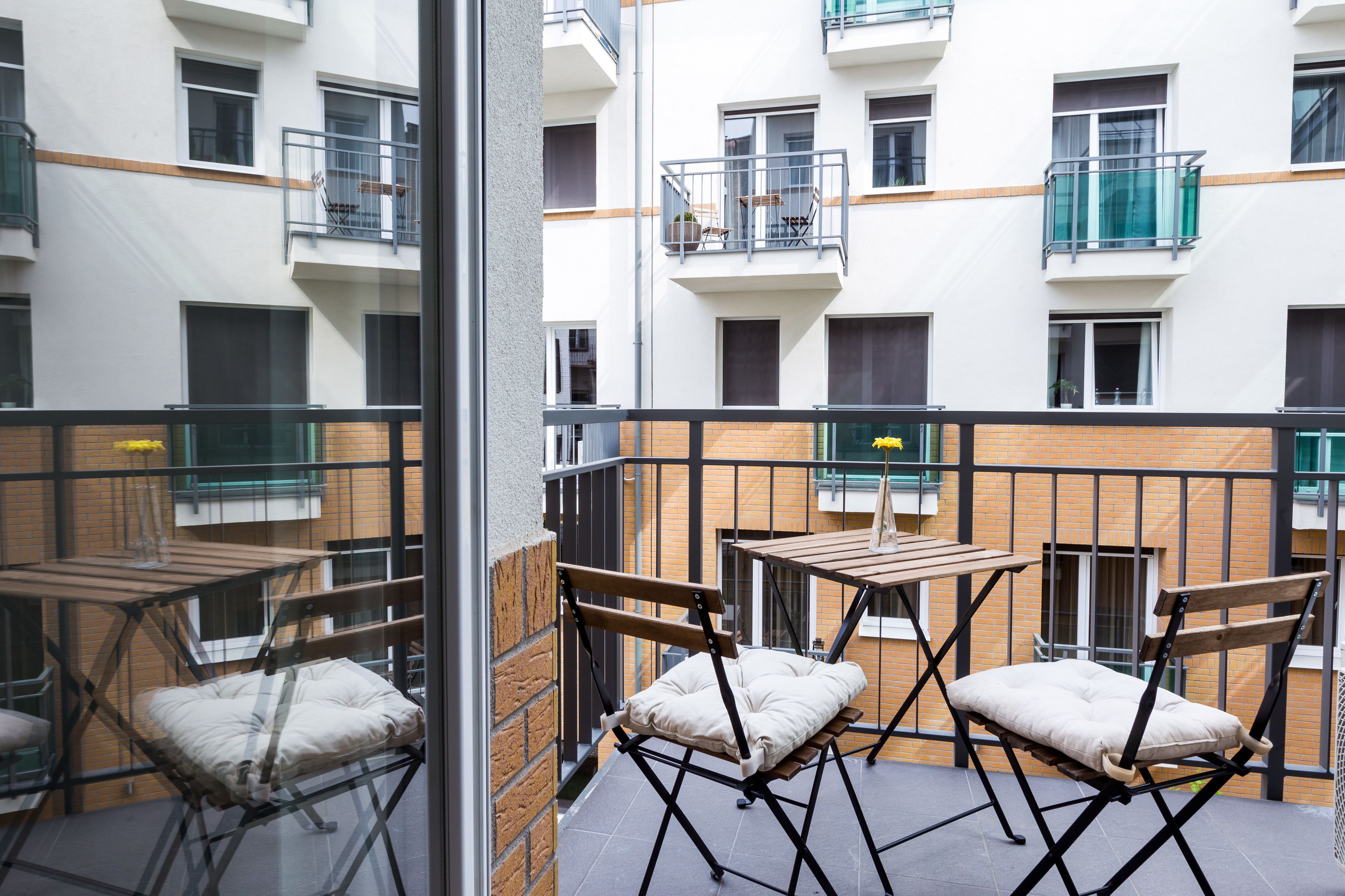 A balcony with a table and chairs, a view of a multi-story building with balconies, and a brick wall.
