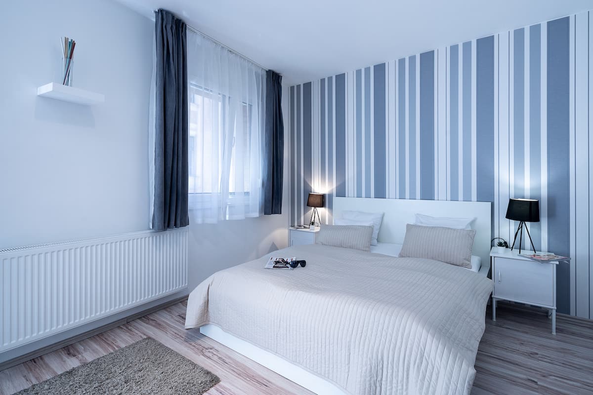 A modern bedroom with a large bed, a bedside table with a lamp, a nightstand, a window with curtains, and a radiator.