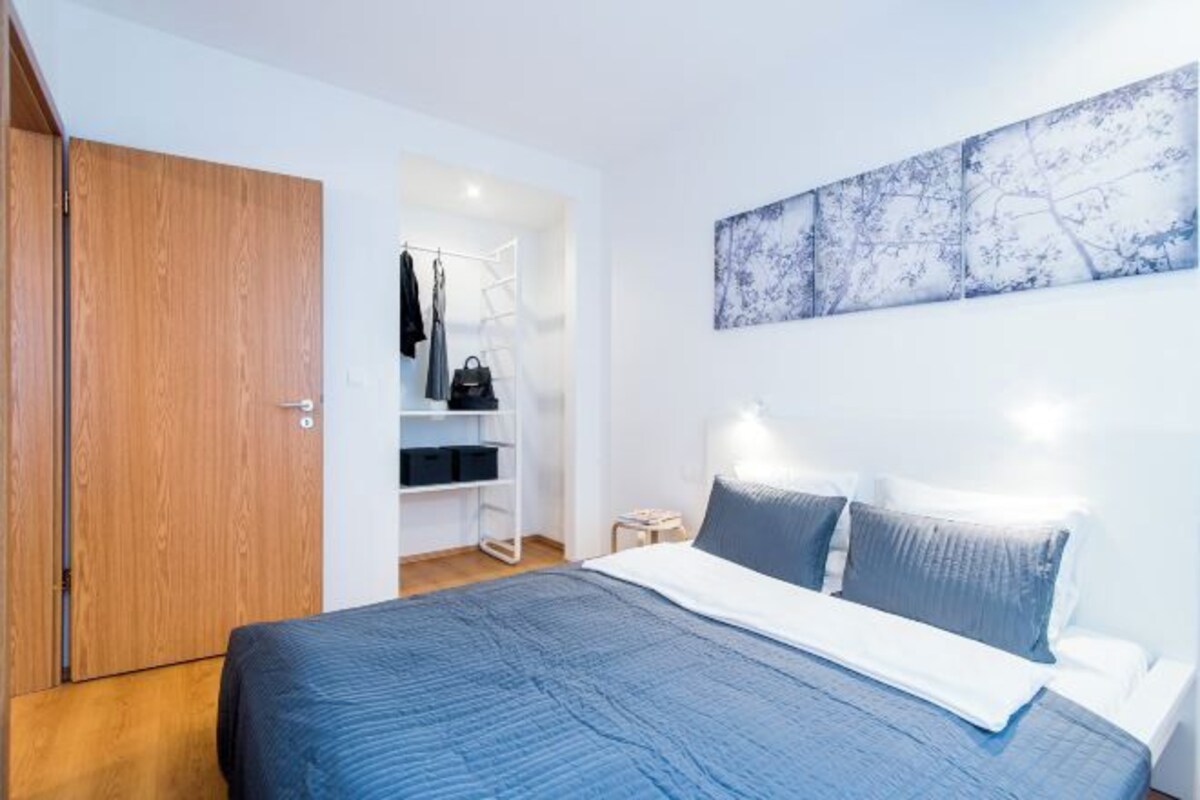 A bedroom with a bed, a wardrobe, a closet with hanging clothes, and a wall-mounted artwork.