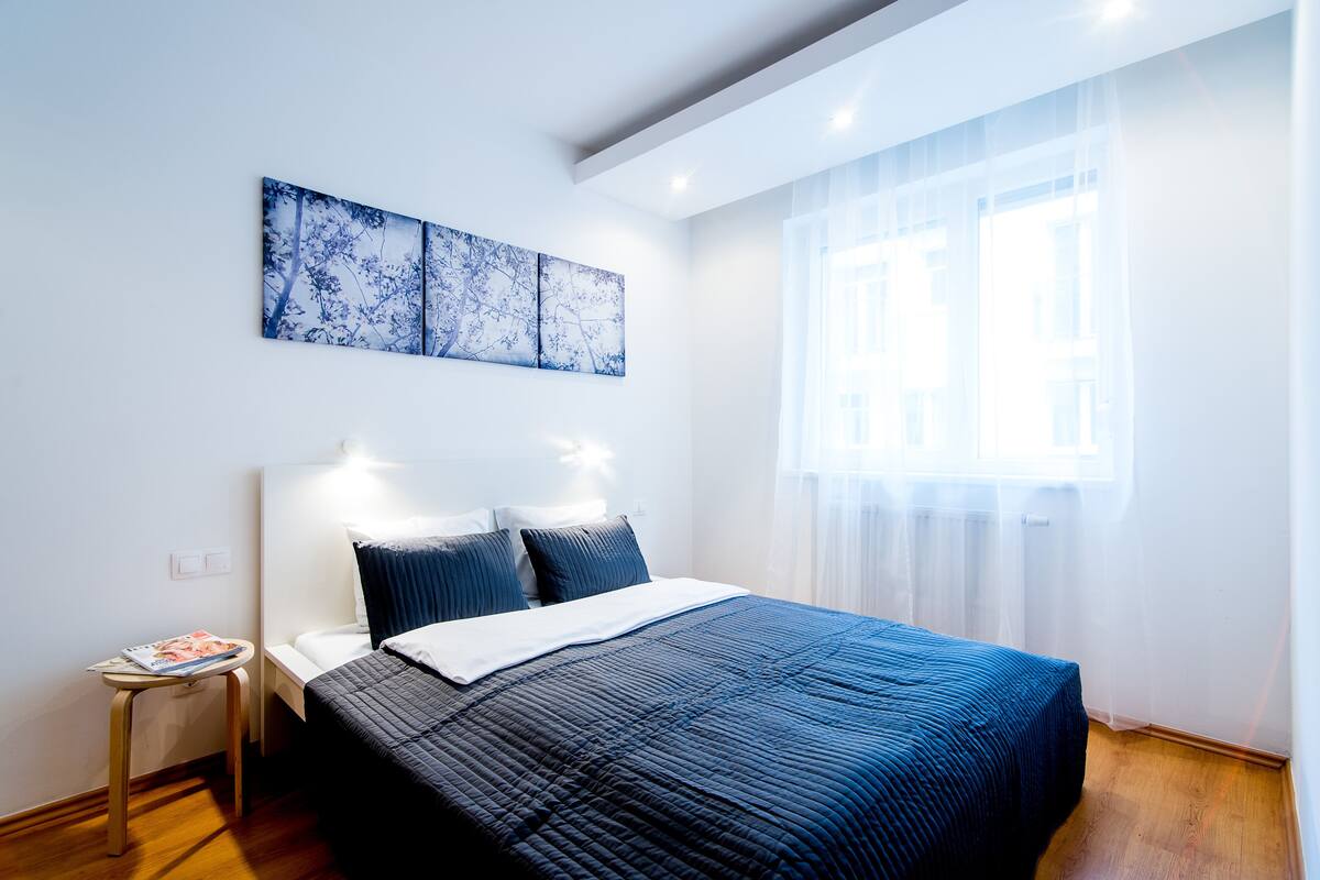 A neatly arranged bedroom with a bed, a small table, and a window with sheer curtains.