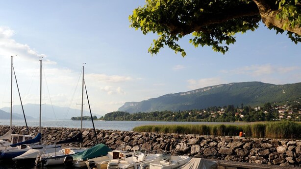 Ein Yachthafen mit vertäuten Booten, eine felsige Küste und der Blick auf eine Stadt am Seeufer, mit Bergen im Hintergrund.