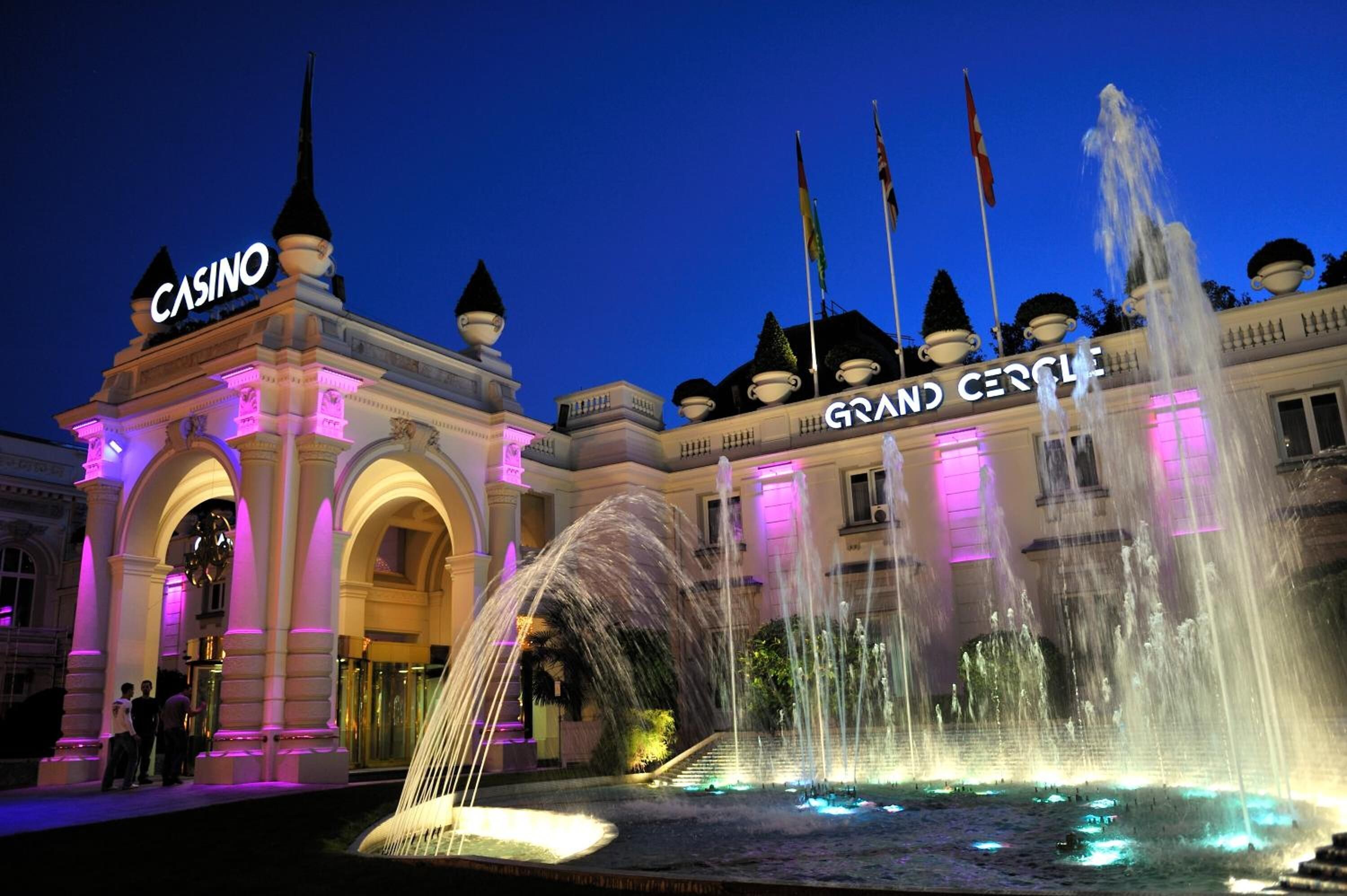 Un imposant bâtiment surmonté d’une grande enseigne indiquant « CASINO » et d’une inscription « GRAND CIRCLE », avec une grande fontaine et une entrée en arc de cercle.