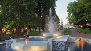 Ein Brunnen, umgeben von einem mit Fliesen ausgekleideten Becken mit Fliesenbordur, Bäumen und einem entfernten Kirchturm.