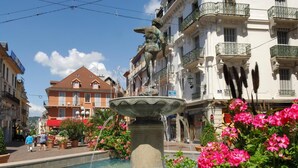 Ein öffentlicher Platz mit einem Brunnen, umgeben von Blumenbeeten und Gebäuden mit Balkonen.