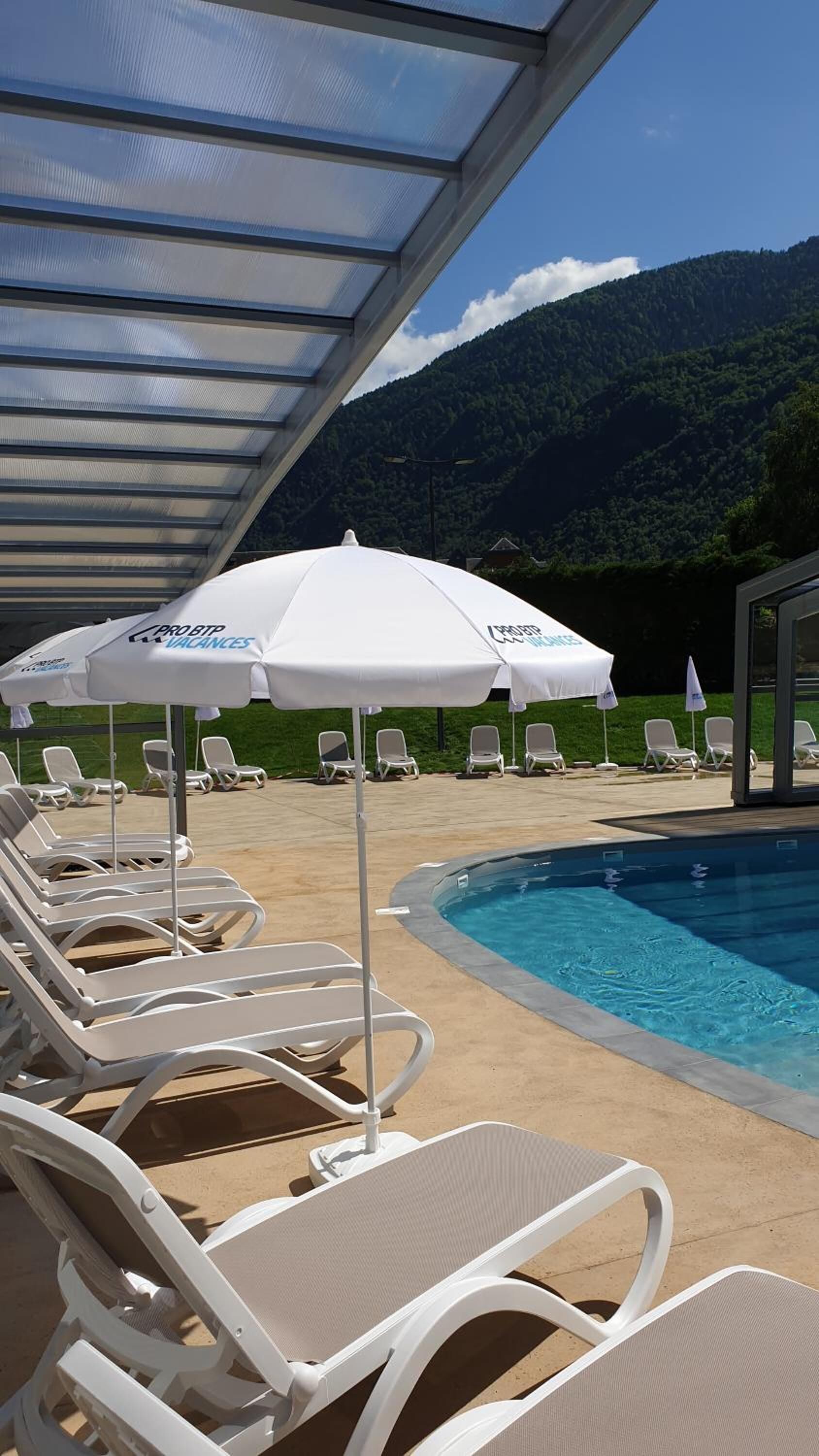 Un espace aménagé au bord de la piscine, avec des chaises longues et des parasols, offrant une vue dégagée sur une chaîne de montagnes, ainsi qu’un espace de détente couvert.