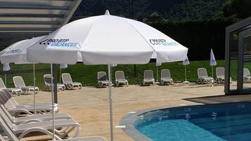 Un espace aménagé au bord de la piscine, avec des chaises longues et des parasols, offrant une vue dégagée sur une chaîne de montagnes, ainsi qu’un espace de détente couvert.