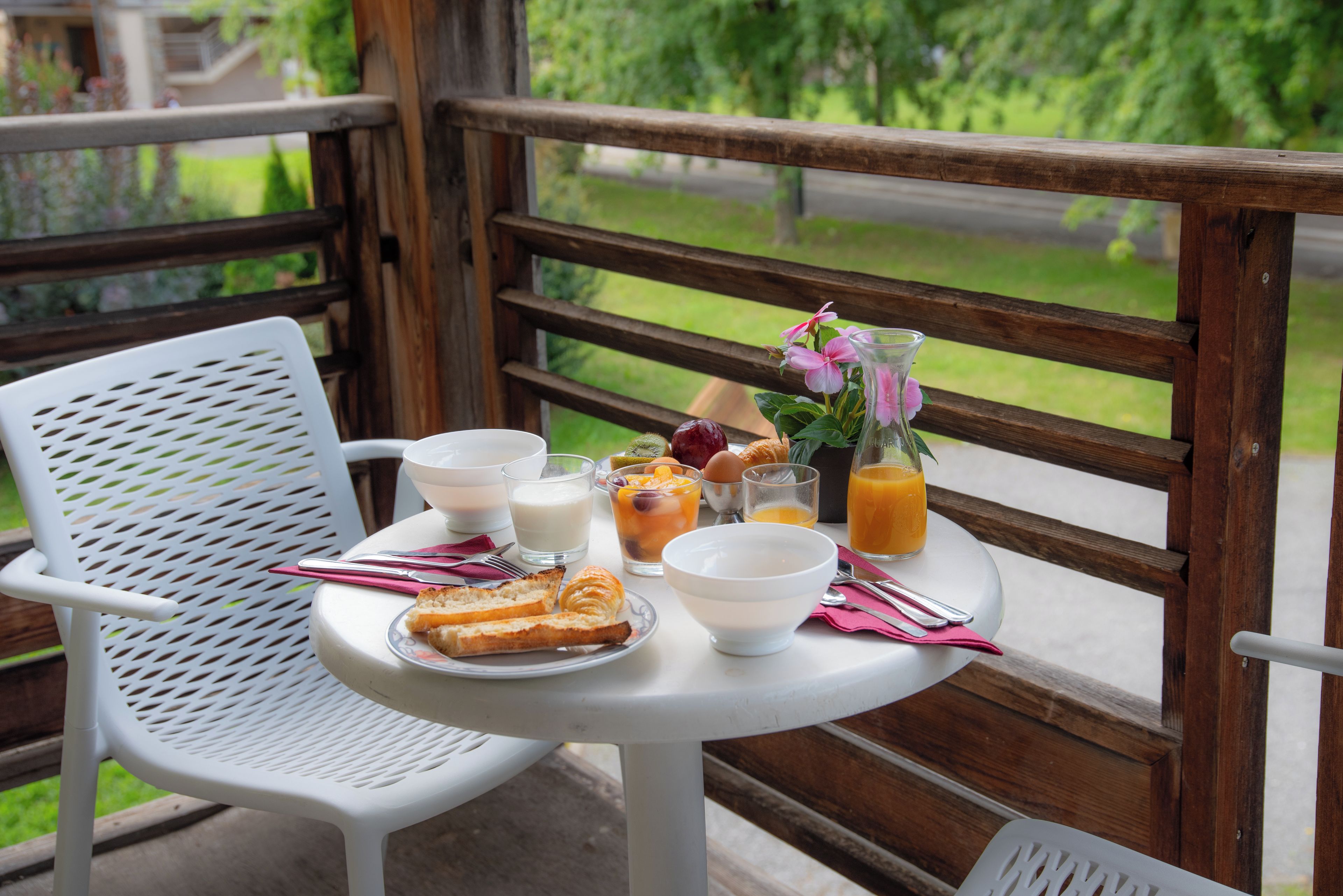 Una mesa con alimentos para el desayuno, como tostadas, huevos y fruta, en un balcón con baranda de madera y follaje al fondo.