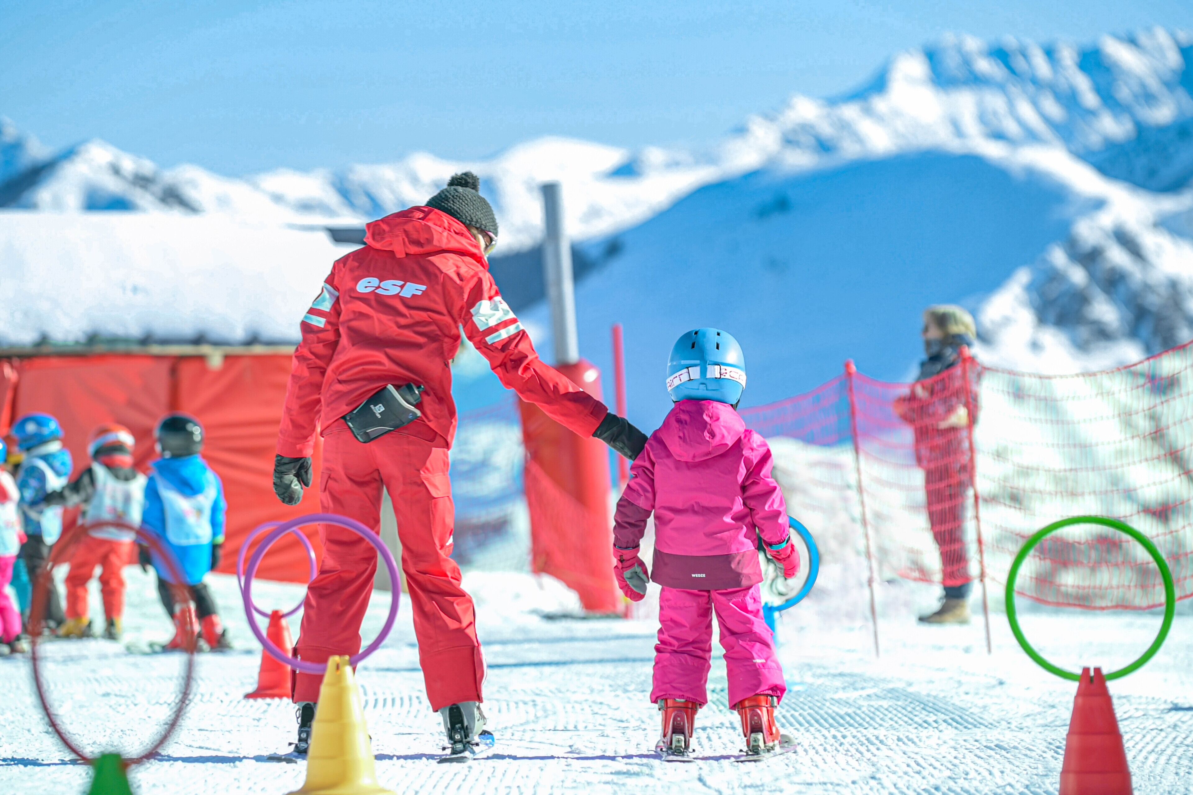 Una pista de esquí nevada con una persona vestida de rojo ayudando a un niño con equipo de esquí rosa.