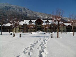 Um hotel com entrada coberta de neve, montanhas ao fundo e céu claro.