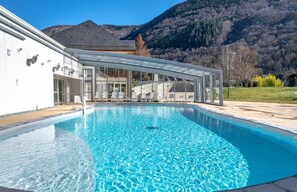 A swimming pool with a glass enclosure, a white building with multiple windows, and a mountainous backdrop. - Village Club Les Balcons des Pyrénées (Saint-Mamet)
