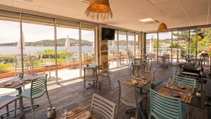 A restaurant with outdoor seating, large windows, and a view of the water. - Village Club Les Mas de L'Esterel (Saint-Raphael)