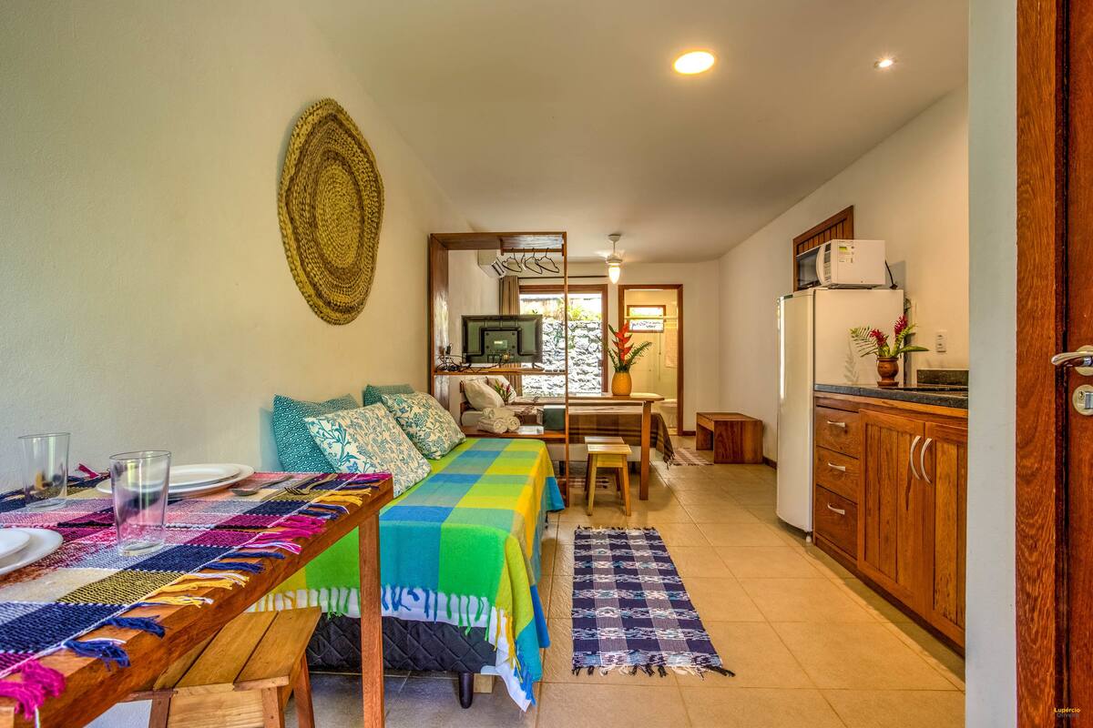 Ein gemütliches Wohnzimmer mit einem Holzesstisch, einer bunten gewebten Tischläufer und einer Couch mit gemusterten Kissen. Im Hintergrund ist eine Küchenzeile mit Holzschränken und einem Kühlschrank zu sehen.