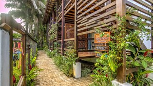 Un sentier mène à une structure en bois sur laquelle est affiché un panneau « Bienvenue », le tout étant entouré d’une végétation luxuriante et de plantes.