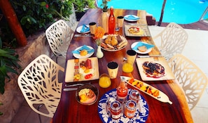 Mesa de jantar com diversos pratos de café da manhã, bebidas e uma piscina ao fundo.