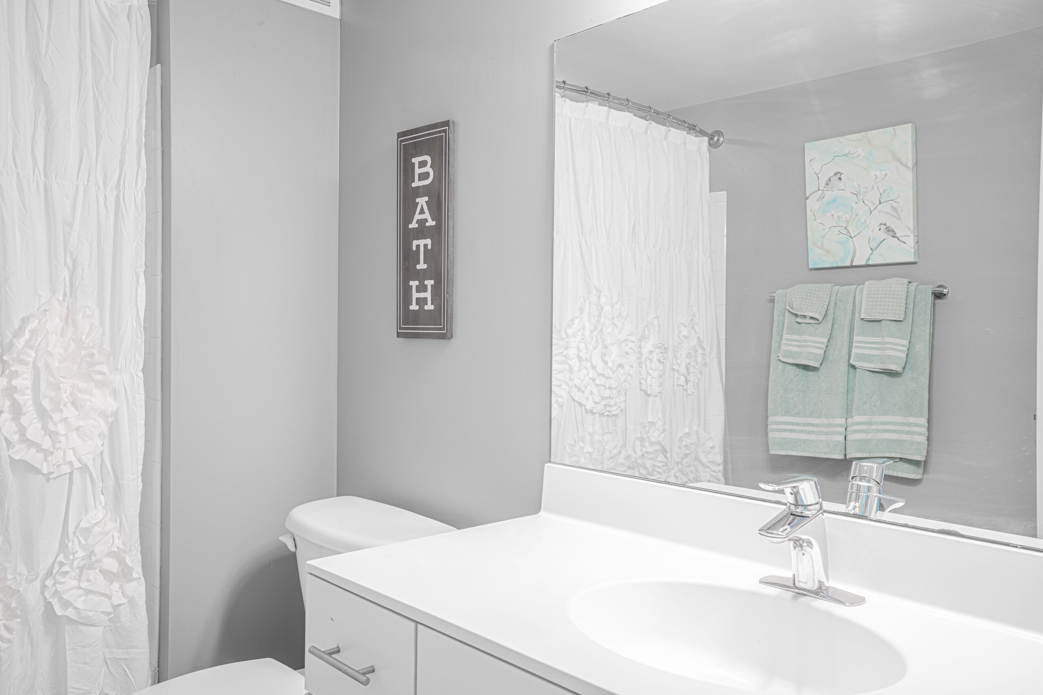 Ein Badezimmer mit einem weißen Waschbecken, einer Toilette, einem Duschvorhang, einem Handtuchhalter mit zwei Handtüchern und einem an der Wand befestigten Schild mit der Aufschrift 'BATH'.