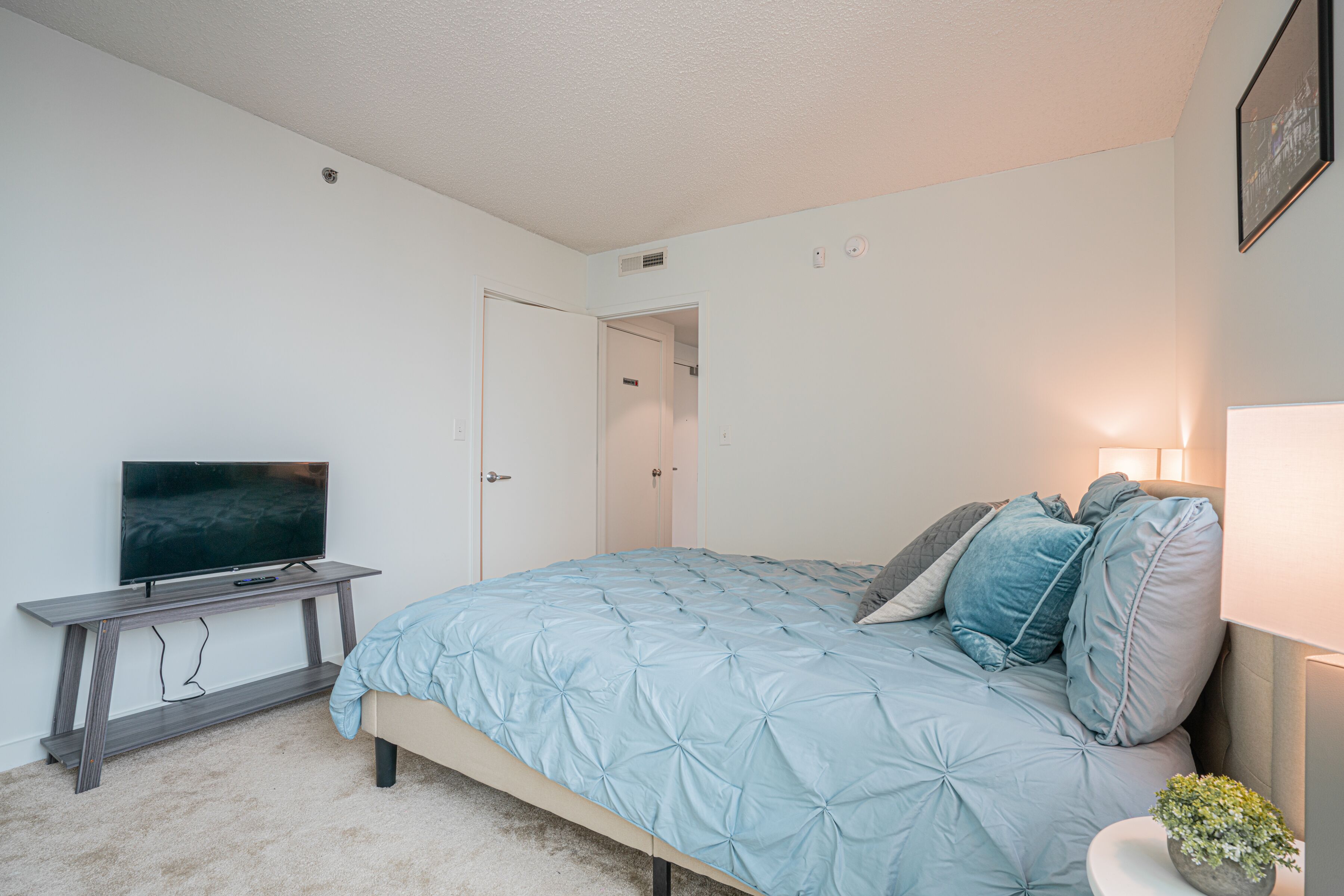 A bedroom with a bed, a TV on a stand, a door, and a wall-mounted light.