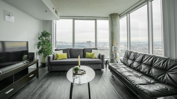 A modern living room with a large window, a grey sofa, a black leather couch, a glass coffee table, and a flat-screen TV. - Heaven on Baltimore Downtown Fully Furnished Apartments (Baltimore)