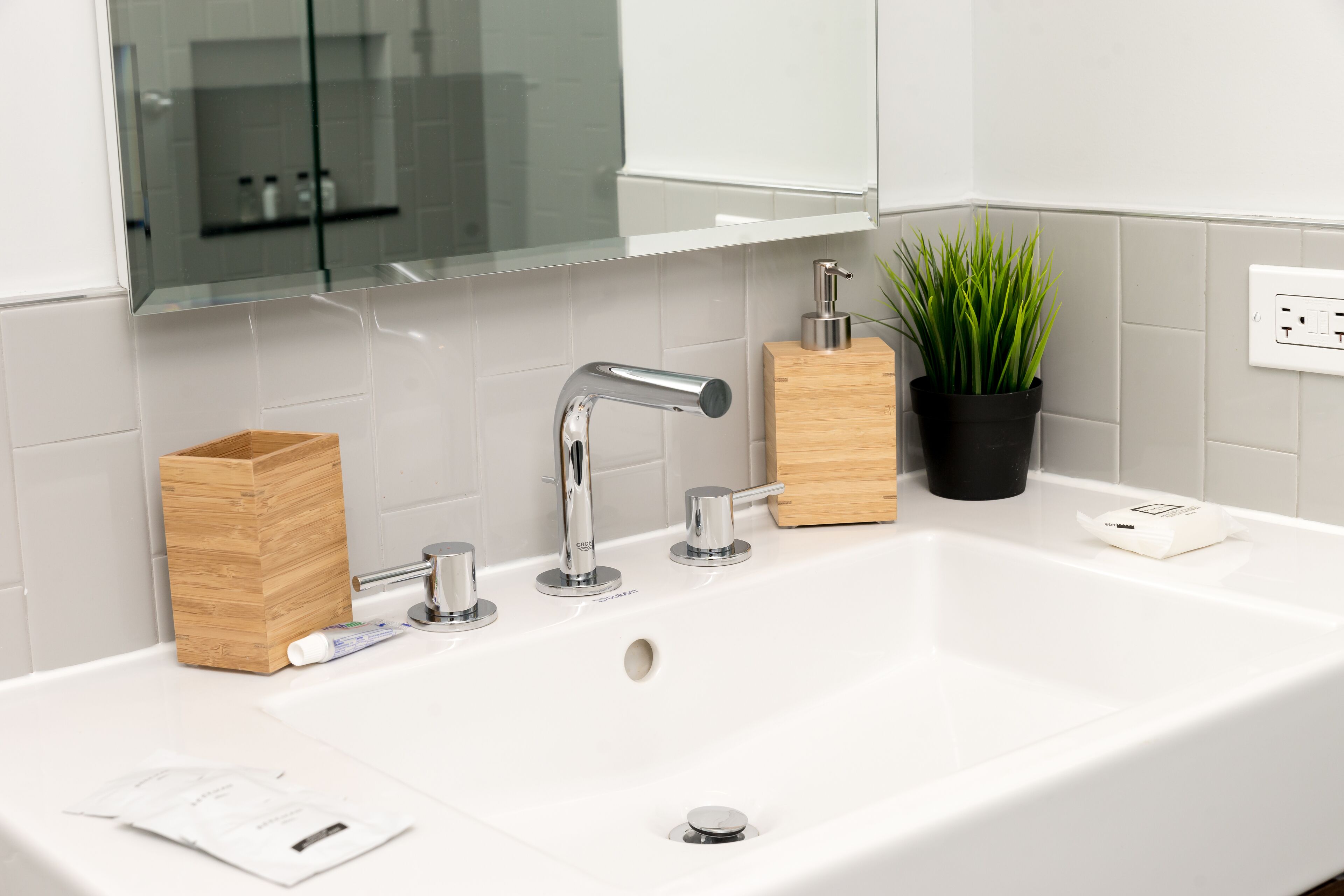 A modern bathroom with a white sink, chrome faucets, a wooden soap dish, and a potted plant.