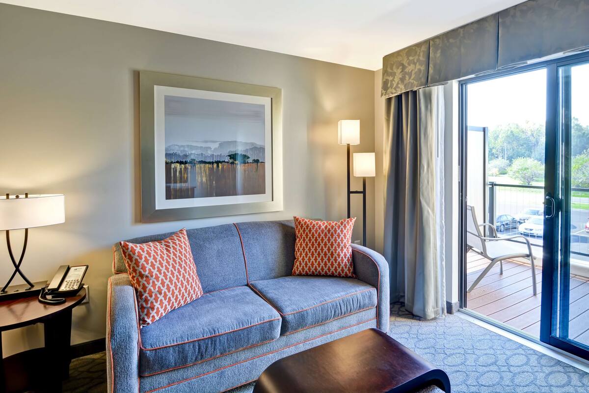 Ein Hotelzimmer mit Sofa, Tisch, Lampe, einem Gemälde und Blick ins Freie.