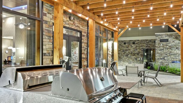 An outdoor kitchen with a stainless steel grill, stone walls, and wooden beams. - Homewood Suites by Hilton New Hartford Utica (New Hartford)