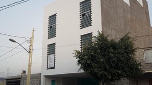 Un edificio moderno de dos pisos con techo plano, varias ventanas con persianas horizontales y un pequeño árbol en la parte delantera.